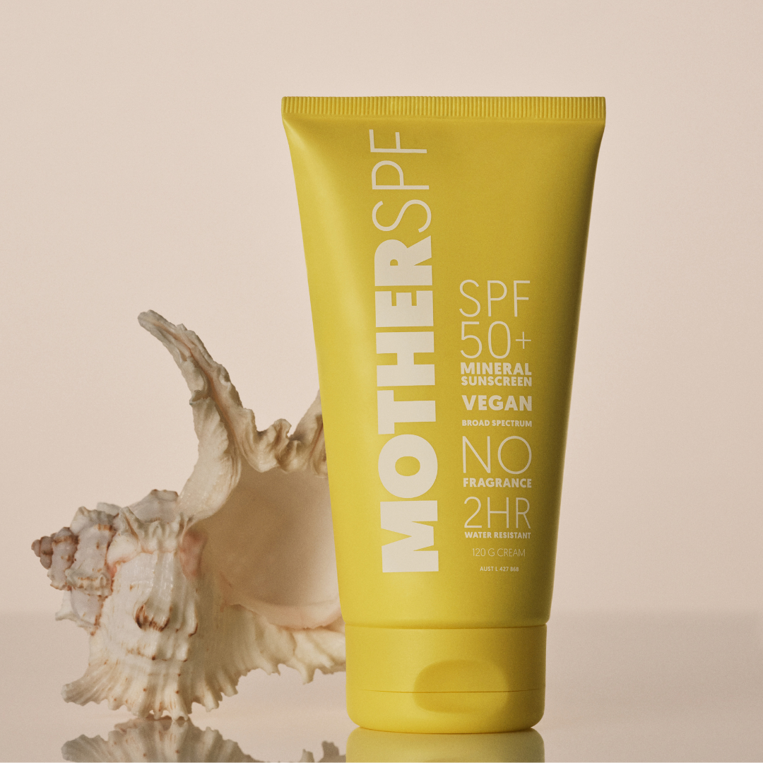 A yellow squeeze tube of MOTHERSPF 50+ mineral sunscreen with white text, standing upright next to a large white seashell on a light beige background.