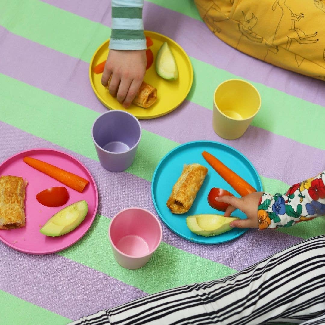 Children's hands reaching for food on colorful plates and cups by bobo&boo on a patterned tablecloth.