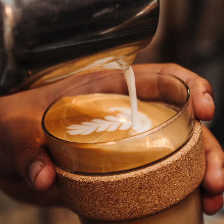 Person pouring milk into a glass of bun coffee with latte art, held by hands.