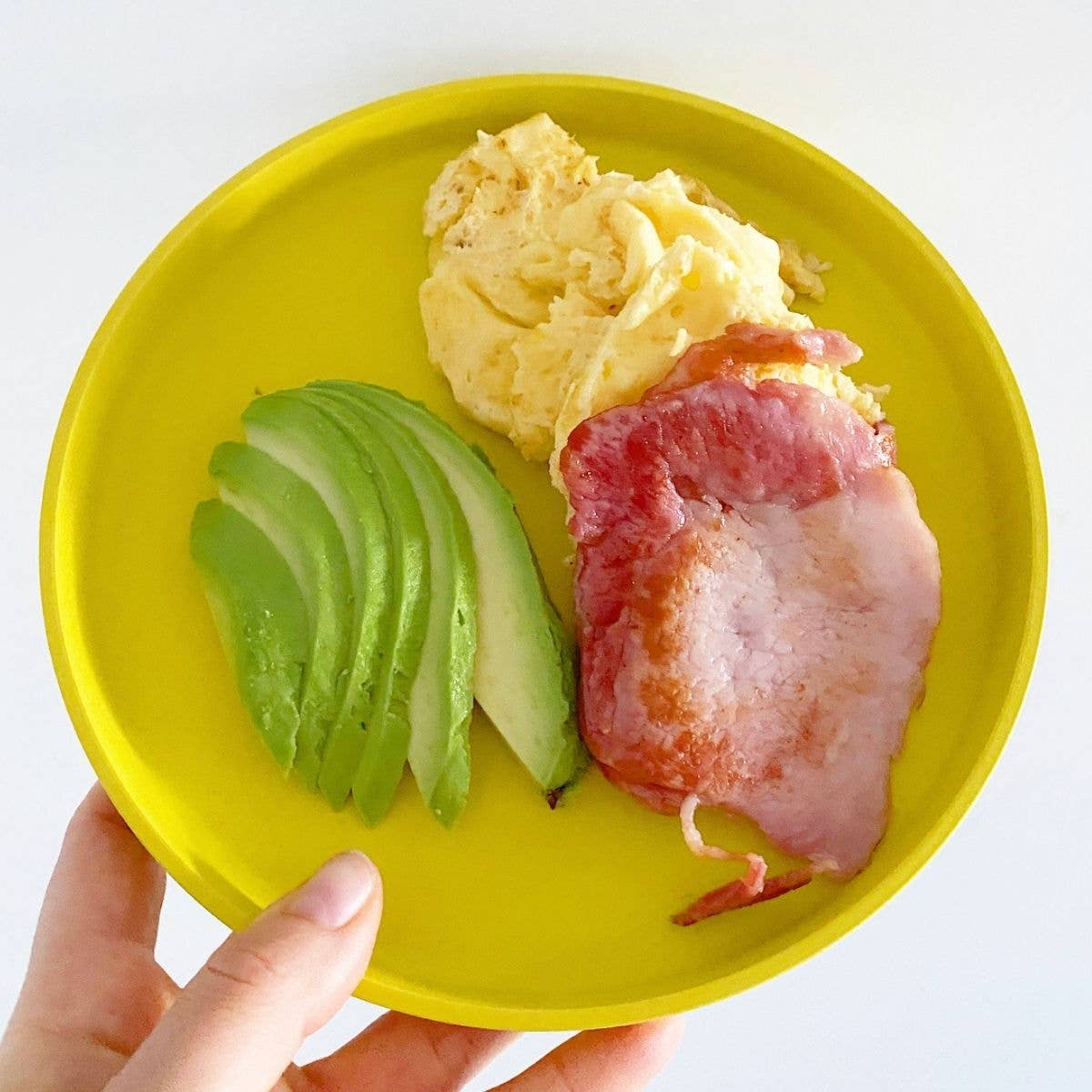 bobo&boo Yellow plate with avocado, scrambled eggs, and bacon held by a hand on a white background