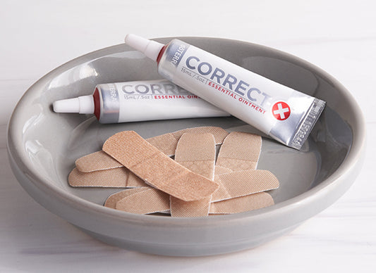 Two tubes of doterra correct x Essential Ointment resting in a gray ceramic bowl alongside several beige adhesive bandages on a light white background.