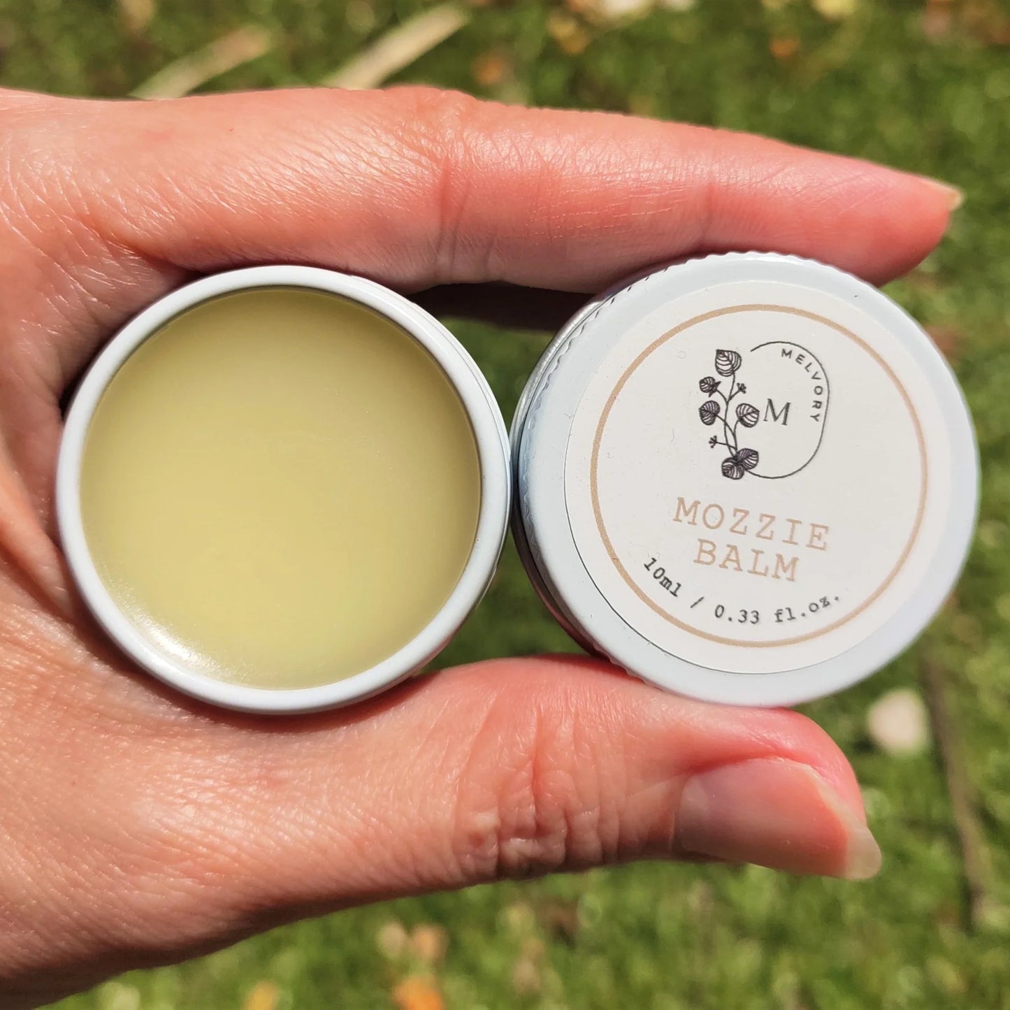Hand holding a tin of melvory Mozzie Balm with a visible brand logo against a natural background.