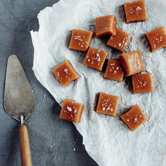 Square caramel candies topped with tasman sea salt flakes, arranged on white parchment paper next to a metal cake server on a dark gray textured surface.