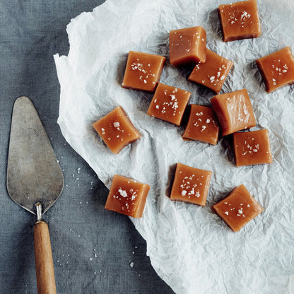 Square caramel candies topped with tasman sea salt flakes, arranged on white parchment paper next to a metal cake server on a dark gray textured surface.