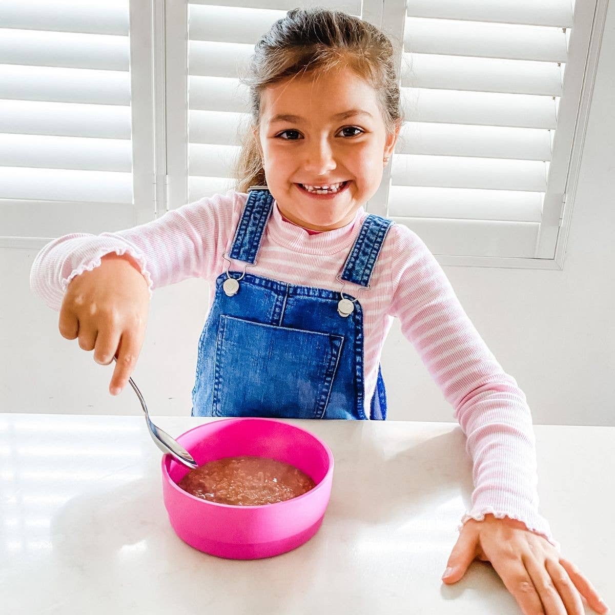 Young girl in a pink shirt and blue overalls pointing at a bobo&boo pink bowl of food on a white table.