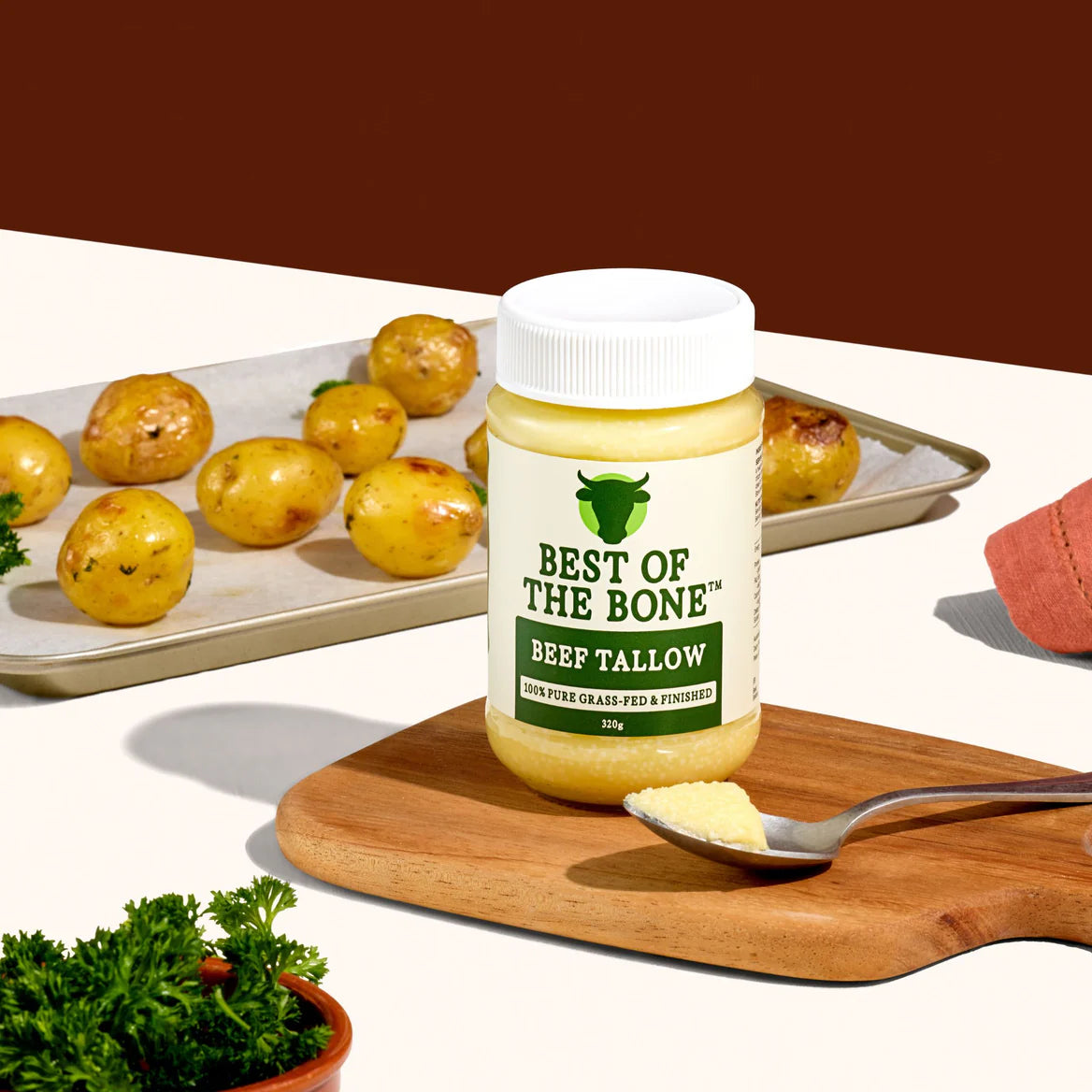 Jar of 'Best of the Bone' beef tallow on a wooden cutting board with a spoonful of tallow and roasted potatoes in the background.