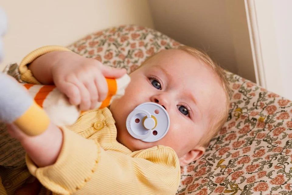 Baby wearing a yellow outfit and orange and white striped socks, sucking bibs dummy