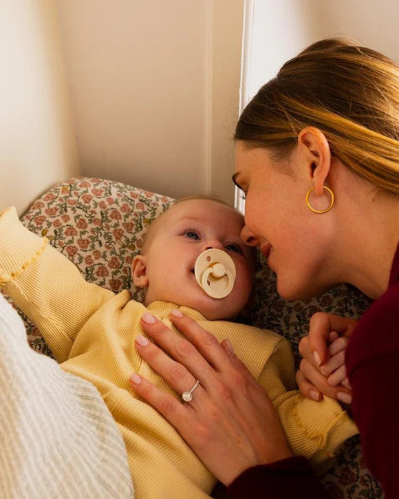 Woman holding a baby wearing a yellow outfit with a bibs pacifier, both looking at each other.