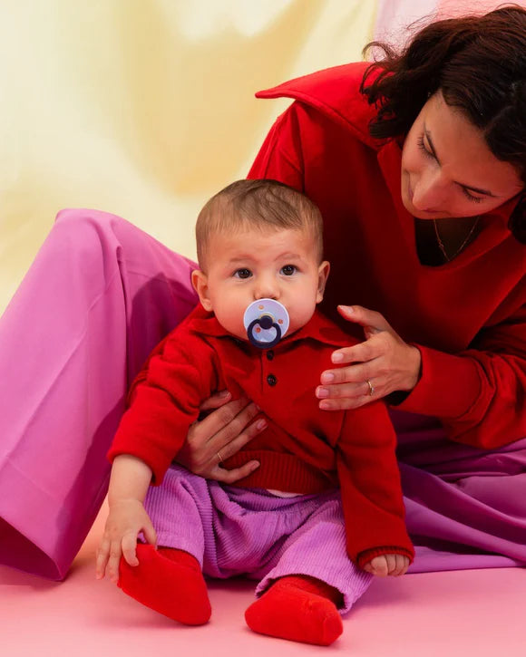 Woman in a red sweater holding a baby in a red outfit with a bibs pacifier, sitting on a pink surface.