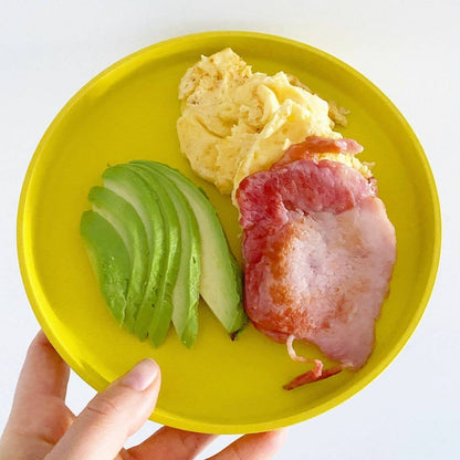 bobo&boo  Yellow plate with avocado, scrambled eggs, and bacon held by a hand on a white background