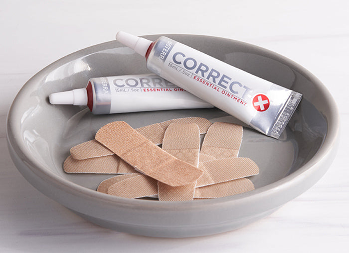 Two tubes of doterra correct x Essential Ointment resting in a gray ceramic bowl alongside several beige adhesive bandages on a light white background.