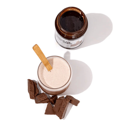 Glass of gevity rx chocolate bone broth with a wooden spoon, jar of chocolate bone broth, and chocolate pieces on a white background