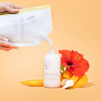 A hand pours melvory mango ad argan oil conditioner from a refill pouch into a white bottle, styled with a red hibiscus flower and yellow mango slices on a warm peach background.