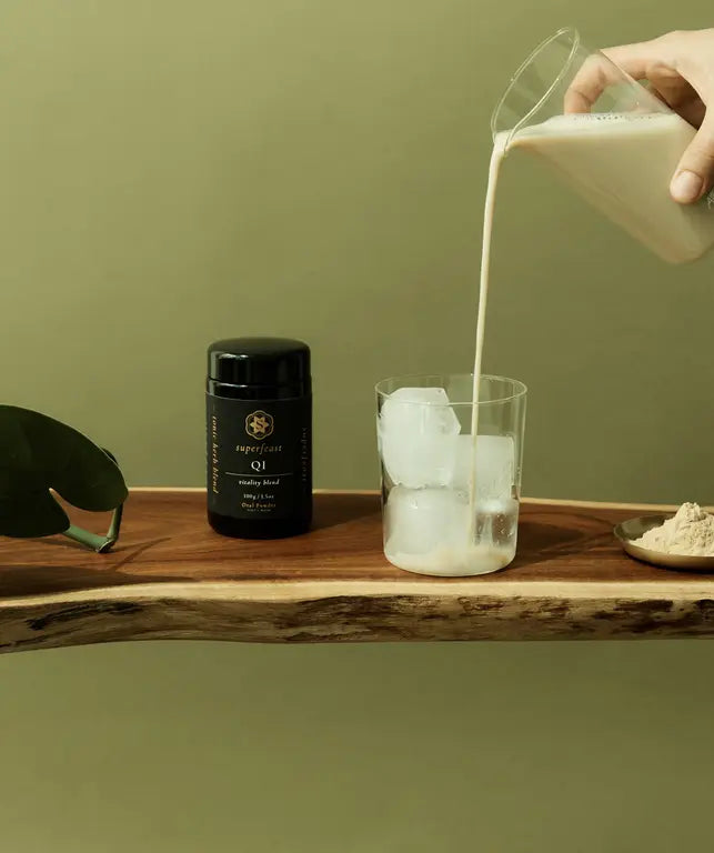 Person pouring milk into a glass with ice cubes on a wooden tray against a green background with a jar of superfeast QI